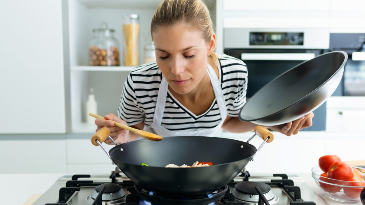 Healthy young woman cooking and smelling food in frying pan in the kitchen at home.