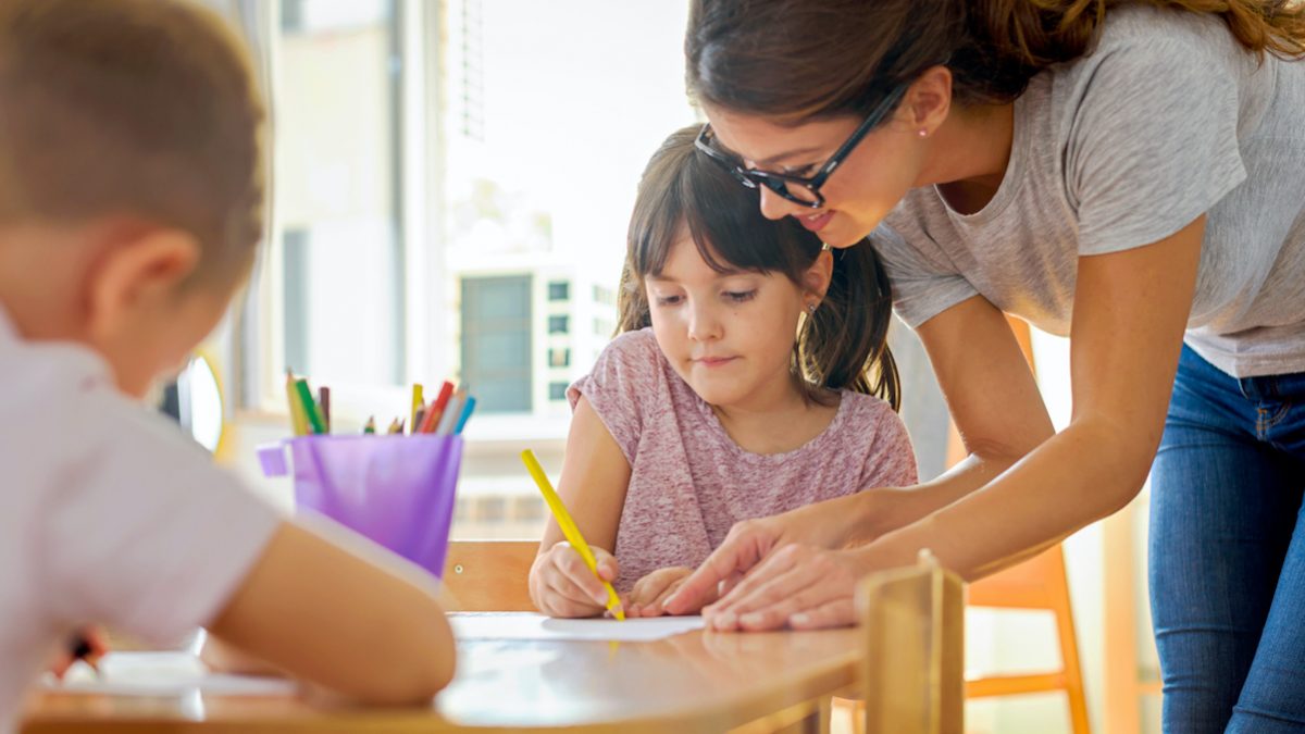 Children drawing with smiling preschool teacher assisting them