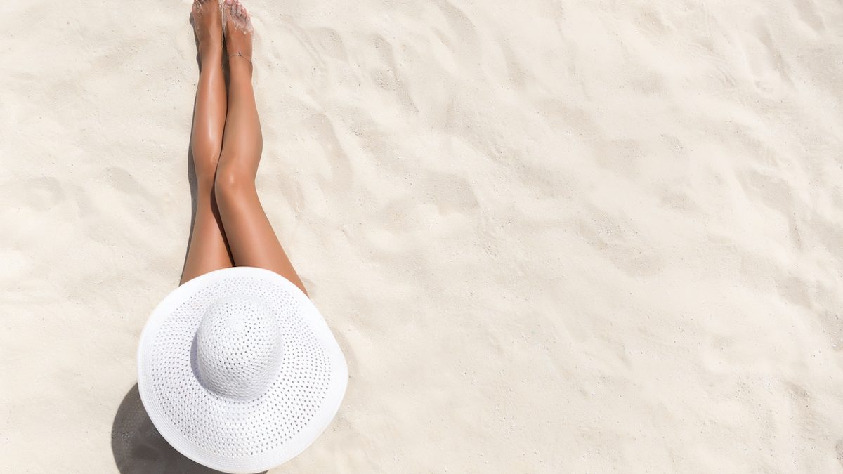 Summer holiday fashion concept - tanning woman wearing sun hat at the beach on a white sand shot from above