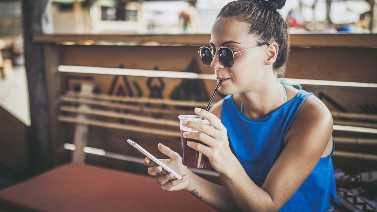 Young woman using mobile phone while drinking juice at beach bar.