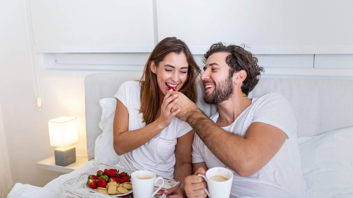 Couple in love having breakfast in bed. Young caucasian couple having romantic breakfast in bed. Female and male , two cups of coffee, fruits and colorful biscuits.